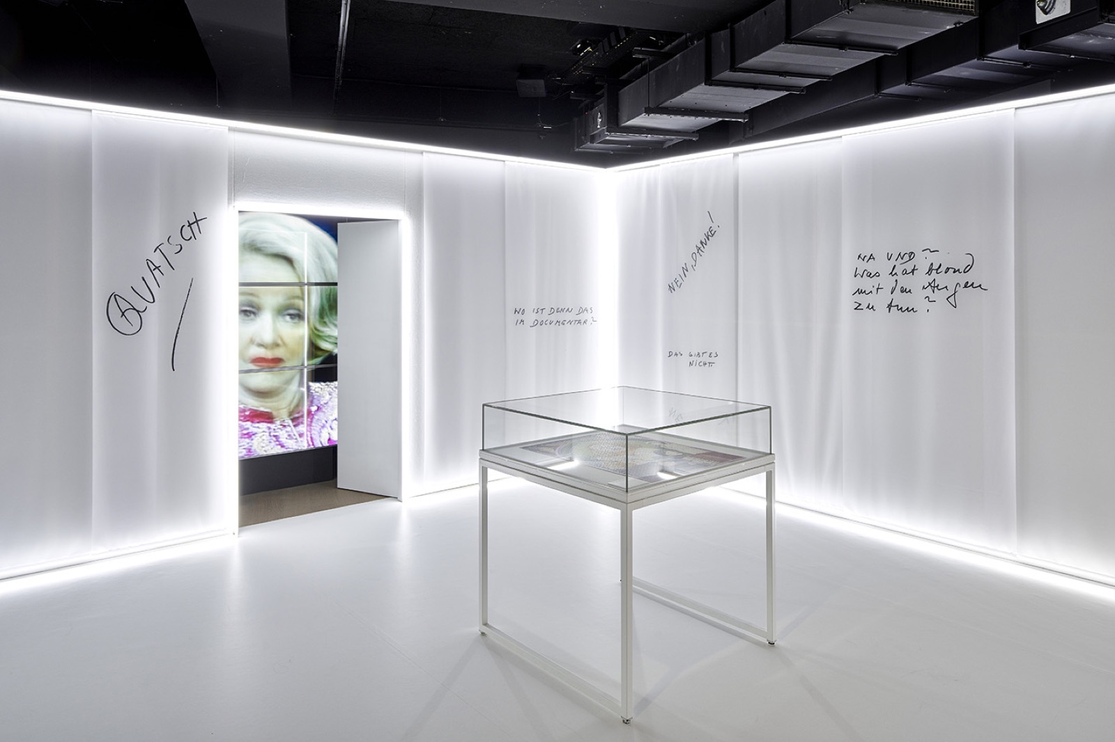 Weißer Raum mit beleuchteten Wänden, handschriftlichen Notizen, einer Vitrine und einem Porträt auf einem Bildschirm im Durchgang