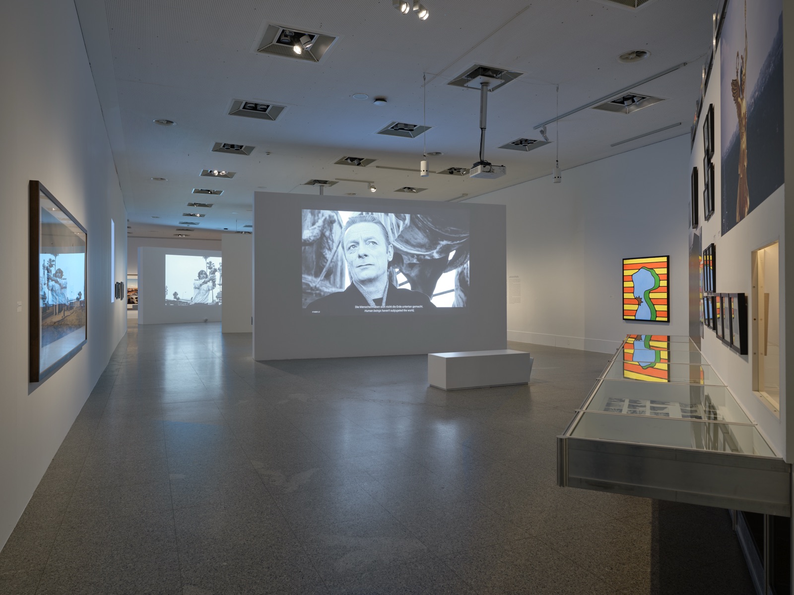 Saal mit einer Projektion, gerahmten Fotografien an der Wand und einer flachen Vitrine